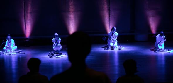 Czrety postacie siedzą w kucki na scenie, są podświetlone na niebiesko. Poza tym scena tonie w ciemności.