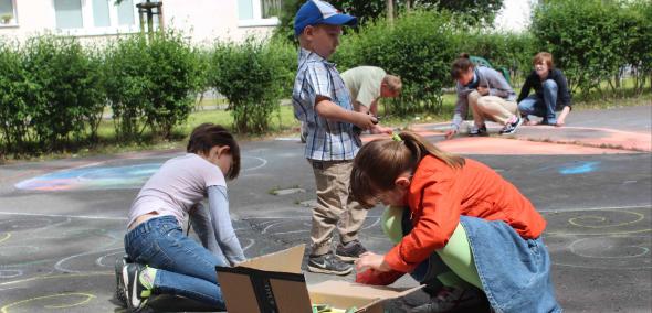 Dzieci bawią się na podwórku wśród budynków. Na asfaltowej nawierzchni malują kolorowymi kredami