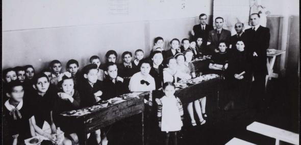 Zdjęcie przedstawia klasę szkolną. W ławkach siedzą dzieci, za nimi w głębi stoi grupa nauczycielek i nauczycieli. Zdjęcie czarno-białe.