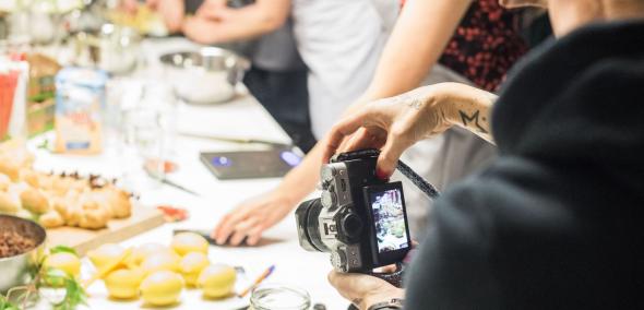 Osoba z aparatem stoi tyłem do widza, na wyświetlaczu aparatu widać kadr fotografii - zastawiony potrawami kolorowy stół, który widać także poza wyświetlaczem aparatu
