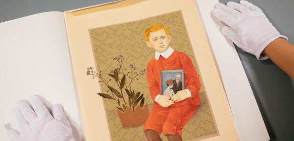 Osoba w białych rękawiczkach ogląda obraz przedstawiający króla Maciusia -rudego chłopca w czerwonej marynarce i czerwonych spodniach do połowy nóg, trzymającego przy pasie, okładką do widza, książkę. Na lewo od chłopca - niebieskie kwiatki, wystające z brązowej schematycznej korony.