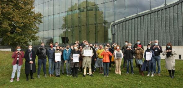 Grupa osób w jesiennych ubraniach stoi przed budynkiem Muzeum POLIN. Część trzyma transparenty, część dłonie złożone w symbol serca. Apel ws. kryzysu uchodźczego na granicy Polski i Białorusi