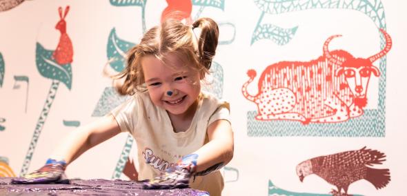 Uśmiechnięta dziewczynka ma ręce umoczone w farbie i maluje na stole. W tle zwierzęta przedstawione w formie kolorowych obrazków w przestrzeni dla dzieci w Muzeum POLIN. 