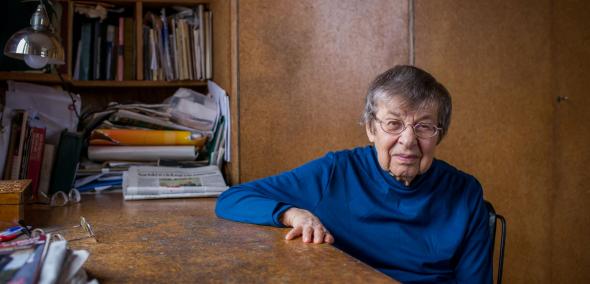 Barbara Góra - starsza kobieta w okularach - siedzi przy stole. Za jej plecami regał pełen książek.
