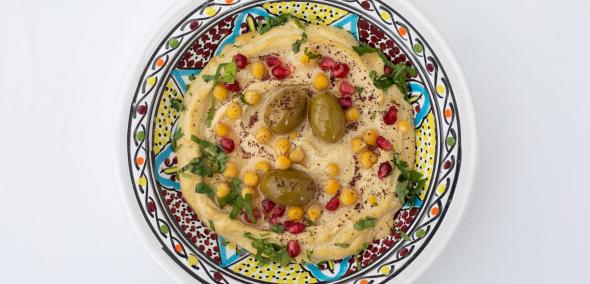 Hummus w kolorowej miseczce. Jest udekorowany oliwkami, ziołami i ciecierzycą.
