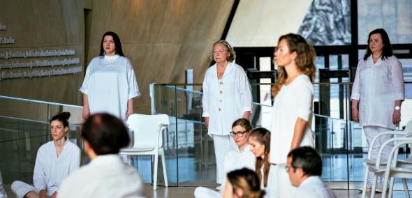 Członkinie chóru POLIN stoją lub siedzą w holu głównym Muzeum POLIN podczas występu.