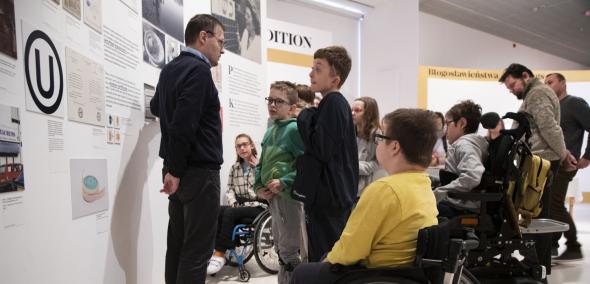 Dzieci z niepełnosprawnościami, w tym na wózkach inwalidzkich, zwiedzają wystawę "Od kuchni" z przewodnikiem.