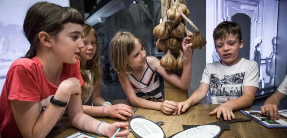 Dzieci siedzą przy stole na wystawie stałej w Muzeum POLIN. Dziewczynka dotyka sztucznego czosnku.