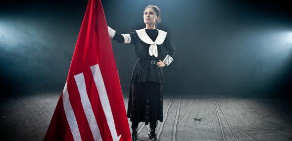 Kadr z filmu "Polskie burki". Kobieta w czarnej sukience z białymi elementami trzyma czerwoną flagę, na której widnieją trzy białe strzałki skierowane do dołu.