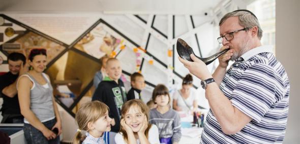 Zajęcia edukacyjne w pawilonie "Muzeum na kólkach". Mężczyzna w jarmułce prezentuje dzieciom dźwięk szofaru, czyli tradycyjnego instrumentu używanego w judaizmie.