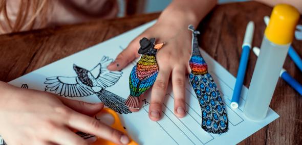 Dziewczynka trzyma dłonie na kartce papieru. Na dwóch palcach ma naklejone kolorowe ptaszki wycięte z papieru.