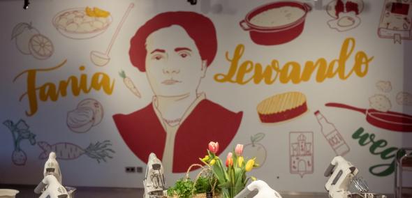 Na stole w pracowni kulinarnej "u Fani" leżą jabłka, stoją garnki i miksery oraz kwiaty w wazonie. W tle mural z podobizną Fani Lewando.