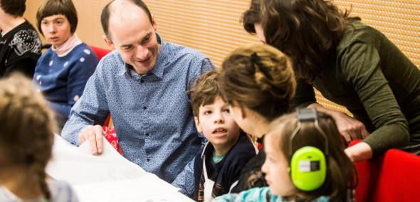 Dzieci z opiekunami siedzą przy stole podczas warsztatów w Muzeum POLIN. Jedna dziewczynka ma założone słuchawki na uszach.
