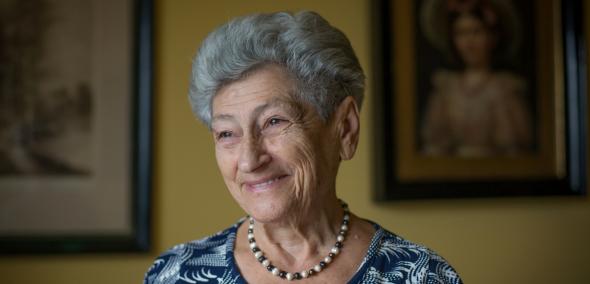 Uśmiechnięta Krystyna Budnicka w jakimś mieszkaniu - starsza kobieta z siwymi włosami, nosi korale na szyi. Jest ubrana we wzorzystą, biało-granatową bluzkę.