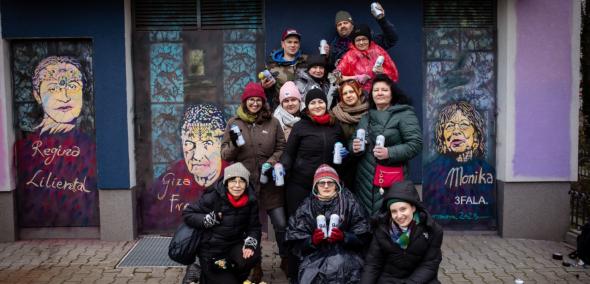 Grupa pracowników Muzeum POLIN trzyma w dłoniach spraye. Za nimi gotowy kolorowy mural.