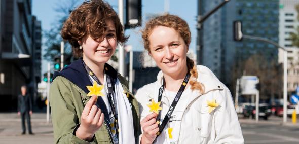 Dwie uśmiechnięte wolontariuszki akcji trzymają przed sobą papierowe żonkile. W tle warszawska ulica.