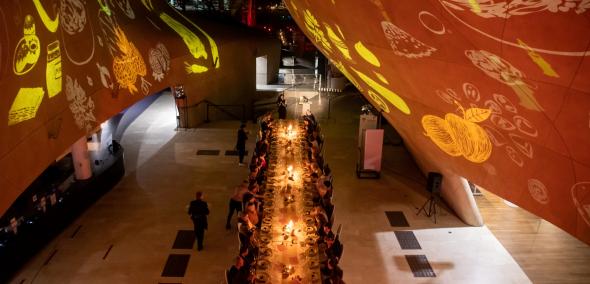 Hol główny Muzeum POLIN. Goście kolacji szabatowej siedzą przy stole. Na ścianie wyświetlają się animacje.