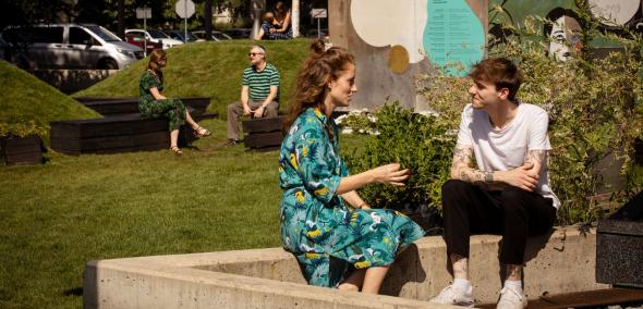 Dziewczyna i chłopak rozmawiają ze sobą, siedząc na murku przy instalacji "POLIN na Łące". W tle, przy muralu siedzą naprzeciw siebie dwie inne osoby.