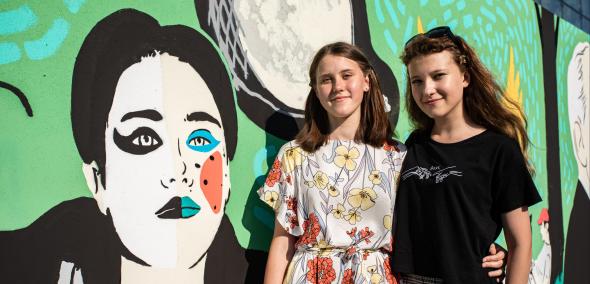 Przy muralu, który przedstawia popiersie młodej osoby z włosami zaczesanymi do tyłu, stoją dwie kobiety, jedna w sukience, druga w czarnej podkoszulce i dżinsach. Obejmują się i uśmiechają.