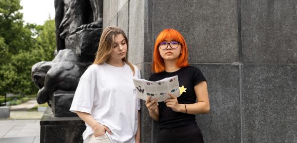 Dwie dziewczyny stoją oparte o pomnik Bohaterów Getta. Dziewczyna z pomarańczowymi włosami trzyma przed sobą ulotkę, na którą spogląda jej brązowowłosa koleżanka.