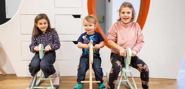 Miejsce edukacji rodzinnej w Muzeum POLIN - dwie starsze dziewczynki i młodszy chłopiec siedzą na konikach na biegunach.