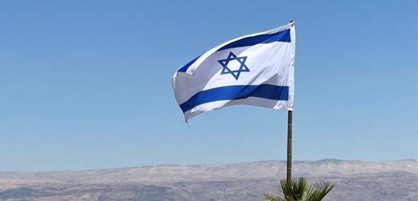 Powiewająca flaga Izraela - biała, z niebieskimi pasami na krawędziach dłuższych boków i granatową Gwiazdą Dawida na środku - w tle wzgórza i błękitne morze