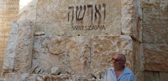 Mur z kamieni w kolorze piasku, z napisami "Warszawa, w języku hebrajskim i polskim. W prawym dolnym roku mężczyzna - Alex Dancyg - w jasnej letniej koszuli.