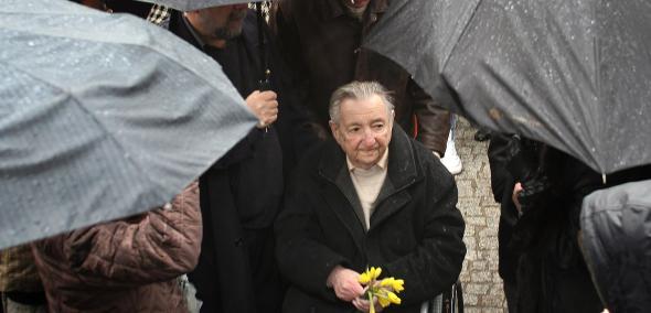 Marek Edelman stoi z odkrytą głową w deszczu. W ręku trzyma kilka żonkili. Wokół niego osoby ukryte pod parasolami. 65. rocznica powstania w getcie warszawskim, przy Kopcu Anielewicza