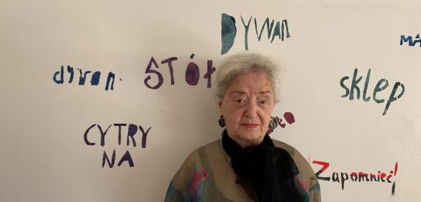 Maya Gordon stoi na tle ściany w Miejskim Ośrodku Sztuki w Gorzowie Wielkopolskim. Na ścianie napisy: cytryna, dzwon, stół, dywan, sklep, zapomnieć.