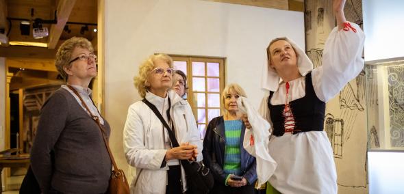 Grupa osób w wieku emerytalnym podczas zwiedzania wystawy stałej Muzeum POLIN. Przewodniczka ubrana jest w historyczny strój karczmarki.