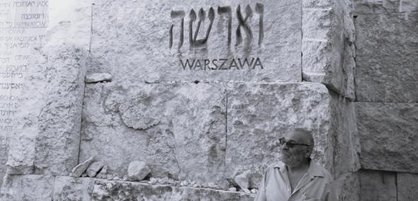 Alex Dancyg stoi przy ścianie z napisem Warszawa.