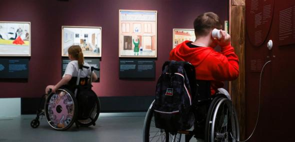 Chłopiec i dziewczyna na wózkach zwiedzają wystawę "(po)ŻYDOWSKIE..." w Muzeum POLIN. Oglądają obrazy Majera Kirszenblata. Chłopiec przykłada słuchawkę do ucha.