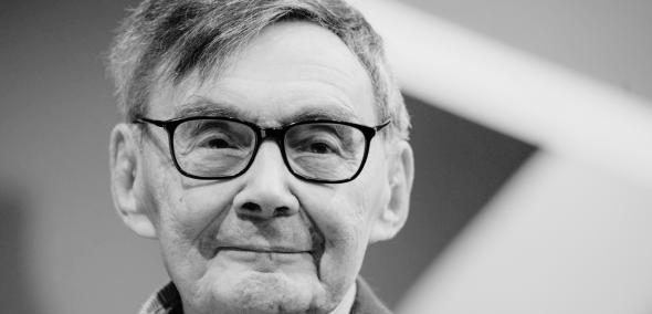 Uśmiechnięty Marian Turski. Zdjęcie w szarej tonacji.