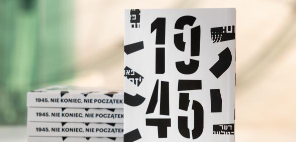 Na stole stoi katalog wystawy "1945. Nie koniec nie początek", obok na stosie leży kilka egzemplarzy publikacji. W tle ściana w holu głównym Muzeum POLIN.