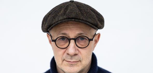 Mikołaj Grynberg - mężczyzna ubrany na czarno, na nosie ma okulary w ciemnych oprawkach, a na głowie szary kaszkiet.