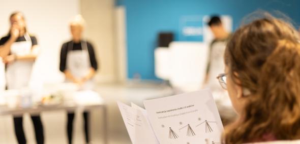 Dziewczyna trzyma przed sobą obrazkową instrukcję zaplatania chałki z sześciu wałków. W tle przy stołach stoją trzy osoby w fartuchach.