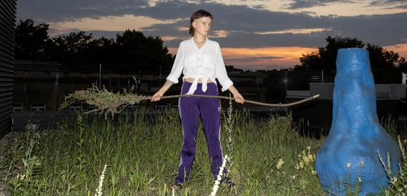 Liliana Zeic stoi na trawie. Trzyma przed sobą gałąź. Obok niej stoi niebieska rzeźba w kształcie nosa.