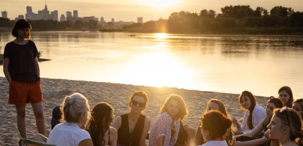 Grupa osób uczestnicząca w Muzealnym Think-tanku siedzi na plaży nad Wisłą w Warszawie.