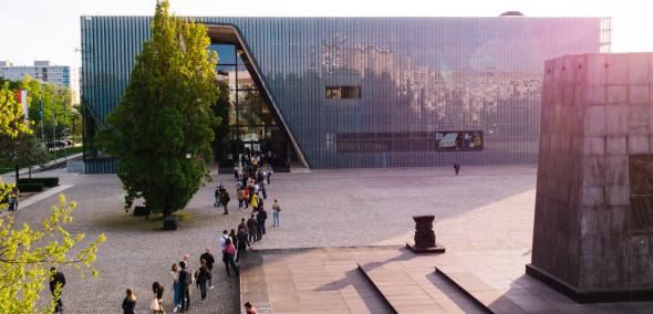 Kolejka osób czeka na wejście do budynku Muzeum POLIN. Świeci słońce. Z prawej strony widoczny pomnik Bohaterów Getta.