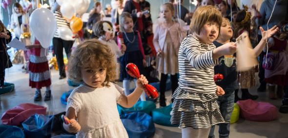 Dzieci z instrumentami muzycznymi tańczą w przestrzeni Muzeum POLIN. Dookoła kolorowe baloniki i inne wesołe ozdoby