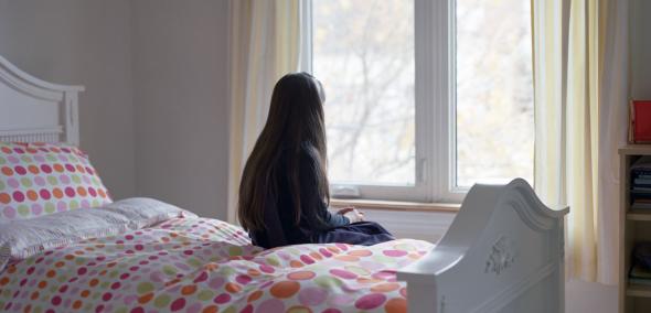 Dziewczyna siedzi na łóżku zwrócona twarzą do okna.