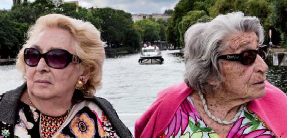 Oma & Bella - film, TISZ Festiwal Żydowskiego Jedzenia - kadr z filmu, przedstawia dwie starsze kobiety, które siedzą obok siebie z głowami odwróconymi od siebie. Każda ma ciemne okulary, narzucony na ramiona sweter oraz barwne bluzki. W tle widok rzeki i miasta położonego na jej brzegach.