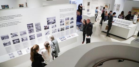 Oprowadzanie w PJM po wystawie "Gdynia - Tel Awiw" - na zdjęciu pojedyncze dorosłe osoby na wystawie, między białymi ścianami działowymi, przyglądają się elementom ekspozycji. Widok z góry.