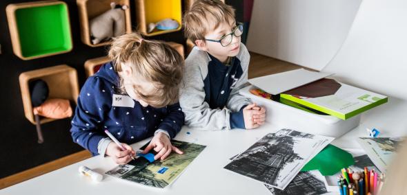 Warsztaty dla szkół w Muzeum POLIN. Na zdjęciu dziewczynka i chłopiec siedzą przy stole nad rozłożonymi starymi zdjęciami i coś do nich doklejają. W tle ściana z kolorowymi półeczkami-boksami.