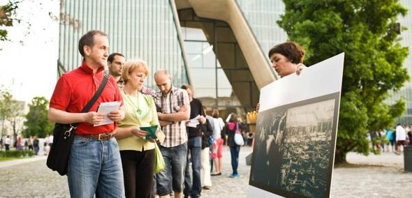 Kilka osób na placu przed budynkiem Muzeum POLIN. Patrzą na obraz, który prezentuje jedna z kobiet.