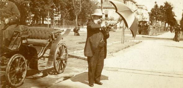 Mężczyzna stoi w parkowej alejce i otwiera parasol. Obok niego na wozie siedzi woźnica.