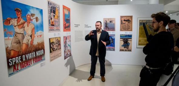 Oprowadzanie kuratorskie po wystawie "Gdynia - Tel Awiw". Na zdjęciu w centralnym miejscu mężczyzna (dr Artur Tanikowski) w garniturze, podaje widocznej po prawej stronie publiczności informacje o plakatach, widocznych na ścianach ekspozycji.