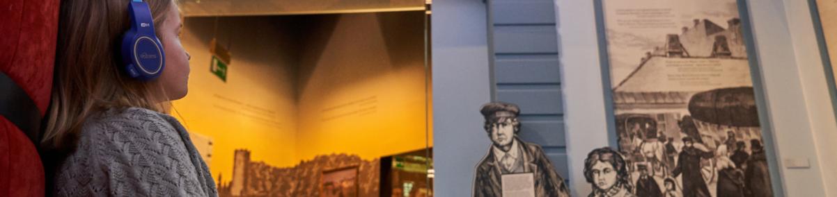 Z lewej strony zdjęcia dziecko, które siedzi na kanapie. W ręku trzyma audioprzewodnik, na uszach ma słuchawki. Patrzy na wystawę w Muzeum POLIN
