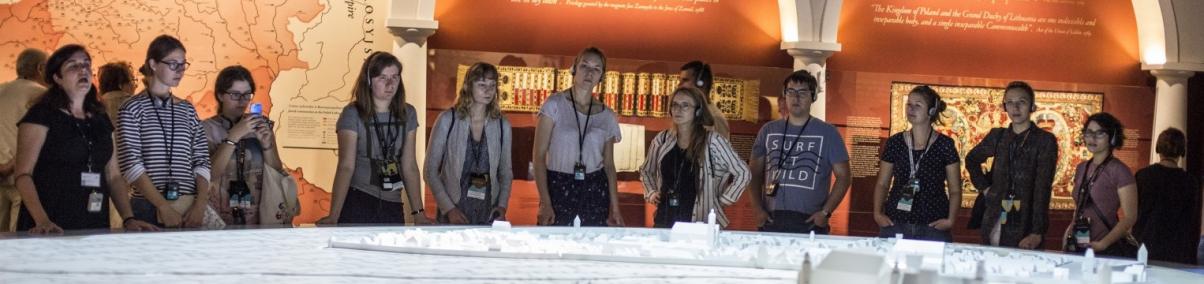 Grupa młodzieży stoi przed ogromną, podświetloną makietą średniowiecznego Krakowa. W tle ściany wystawy "1000 lat historii Żydów polskich" Muzeum POLIN
