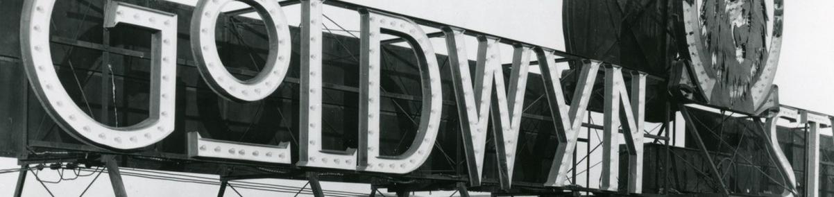 Czarno białe zdjęcie przedstawia ogromny neon "Goldwyn". Pod jego konstrukcją siedzi trzech mężczyzn.